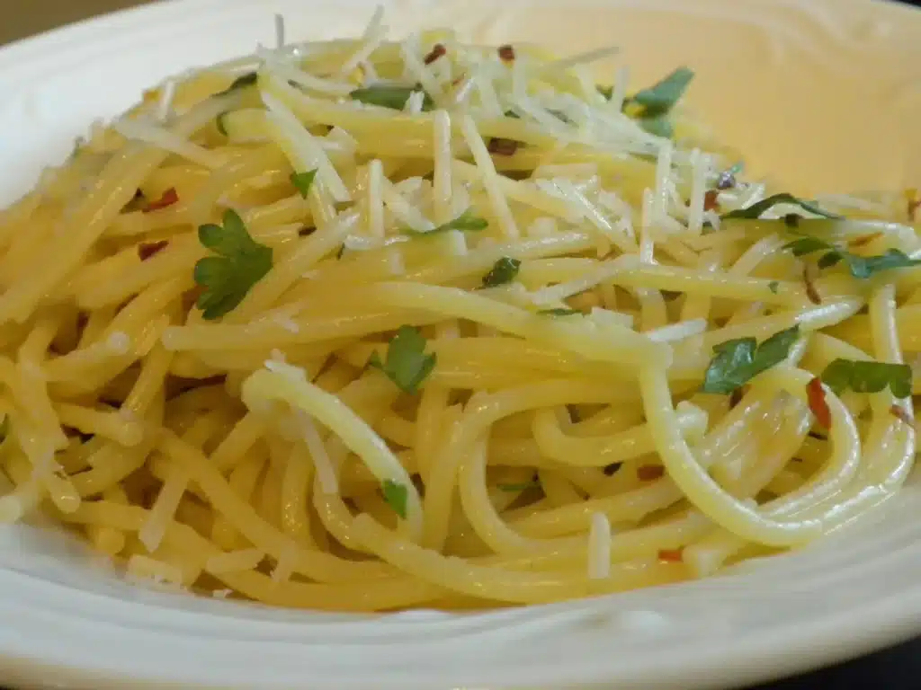 A plate of garlic butter pasta spaghetti topped with grated cheese, chopped parsley, and red pepper flakes on a white dish.