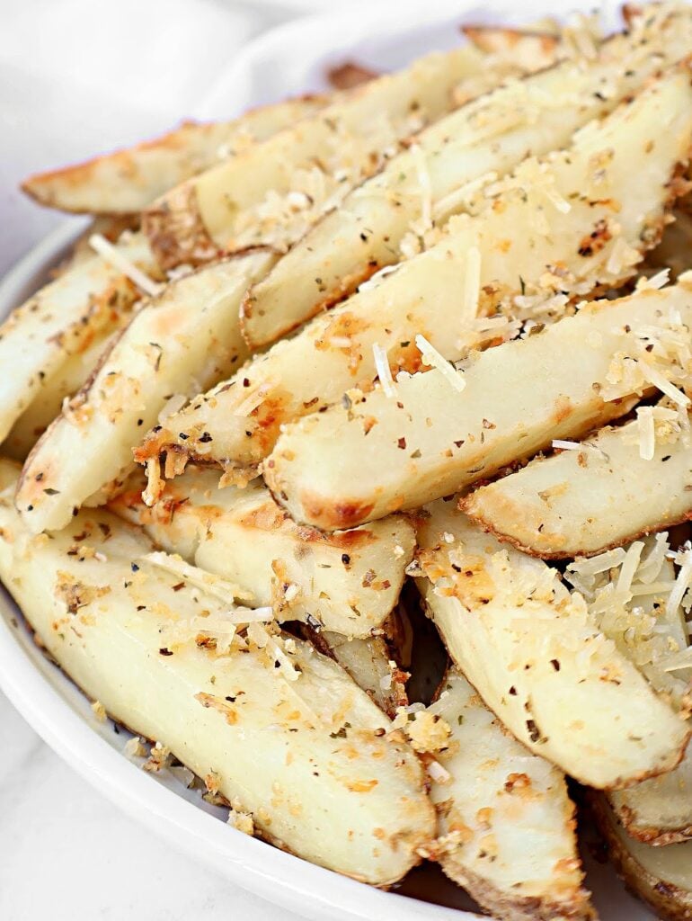 A close-up of a plate piled with homemade parmesan potato fries, seasoned with herbs and topped with grated parmesan cheese. The wedges have golden edges and a rustic appearance, like oven fries with Italian seasoning and skin on.