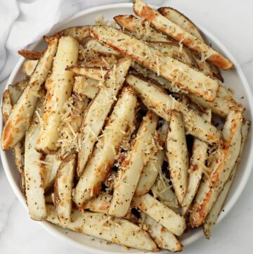 A plate of Parmesan potato fries topped with grated cheese and sprinkled with herbs and seasoning, served on a white background. These oven fries with Italian seasoning offer a savory twist to classic potato wedges.