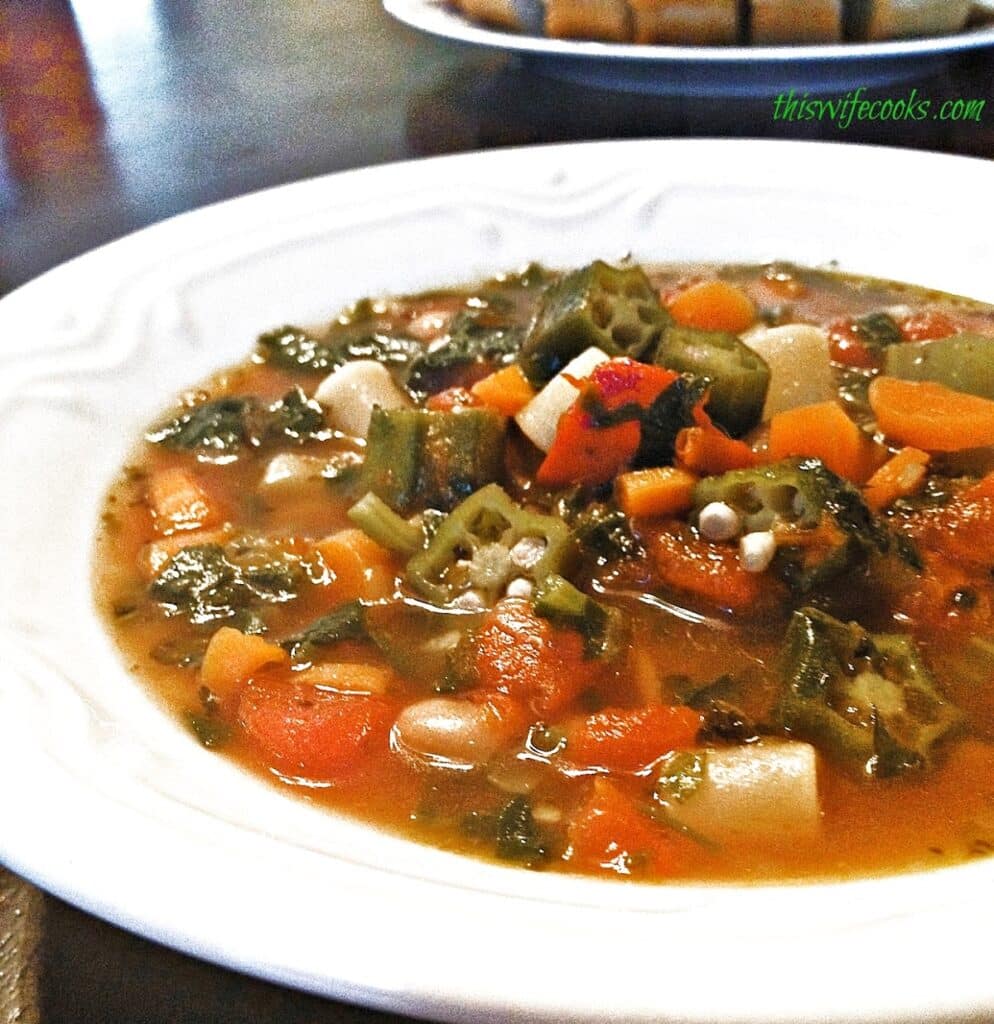 A close-up of a bowl of Late Summer Garden Minestrone, featuring okra, carrots, tomatoes, beans, and greens in a clear broth, served on a white plate.