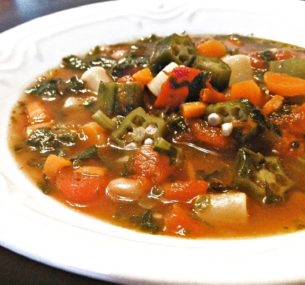 A bowl of Garden Vegetable Minestrone Soup features chopped okra, carrots, tomatoes, white beans, and leafy greens in a savory broth, served in a white bowl.