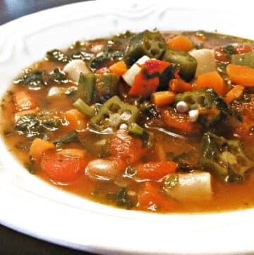 A bowl of Garden Vegetable Minestrone Soup features chopped okra, carrots, tomatoes, white beans, and leafy greens in a savory broth, served in a white bowl.