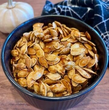 A black bowl filled with sweet and spicy roasted pumpkin seeds sits on a wooden surface. A small white pumpkin and a dark cloth with a white web pattern are in the background.