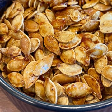 A close-up of sweet and spicy roasted pumpkin seeds coated with seasoning, displayed in a dark bowl. The glossy, brown seeds suggest they are freshly roasted and packed with bold flavor.