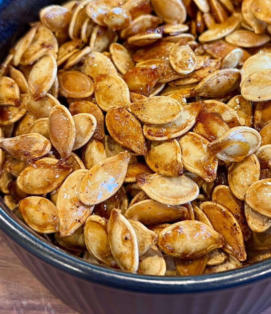 A close-up of sweet and spicy roasted pumpkin seeds coated with seasoning, displayed in a dark bowl. The glossy, brown seeds suggest they are freshly roasted and packed with bold flavor.
