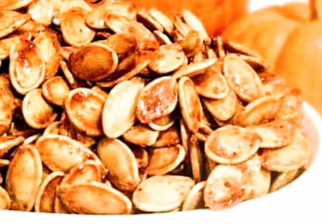 A close-up of a bowl filled with Sweet and Spicy Pumpkin Seeds, seasoned and toasted to a golden brown, with blurred pumpkins visible in the background.