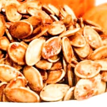 A close-up of a bowl filled with Sweet and Spicy Pumpkin Seeds, seasoned and toasted to a golden brown, with blurred pumpkins visible in the background.