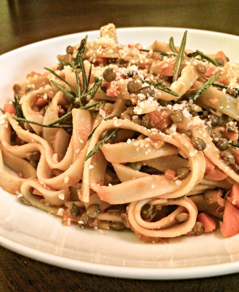 A close-up of Lentil Bolognese pasta garnished with diced carrots, fresh rosemary sprigs, and grated cheese, served on a white dish with a dark background—a perfect vegan pasta sauce option.