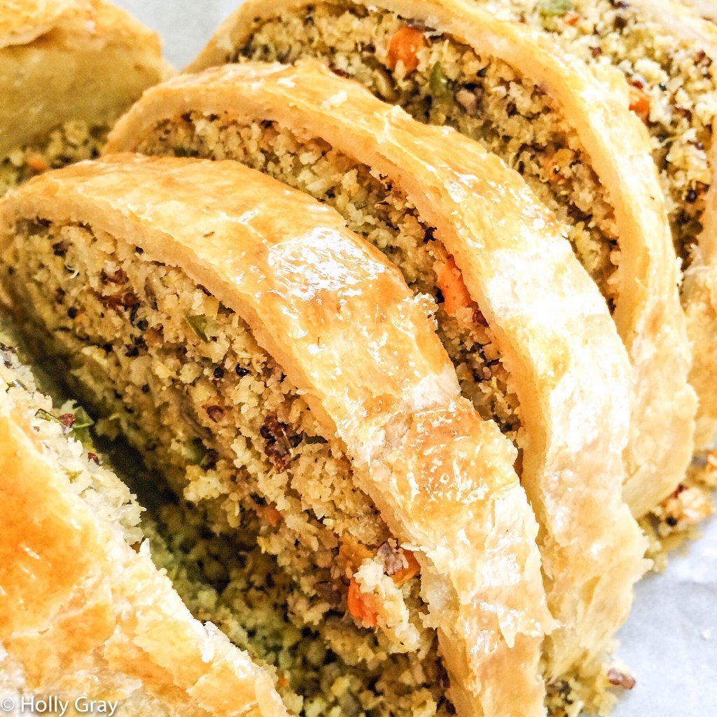 Close-up of sliced vegetable Wellington with a golden, flaky pastry crust and a vibrant filling of grains, vegetables, and herbs. The moist interior shows colorful orange and green specks.