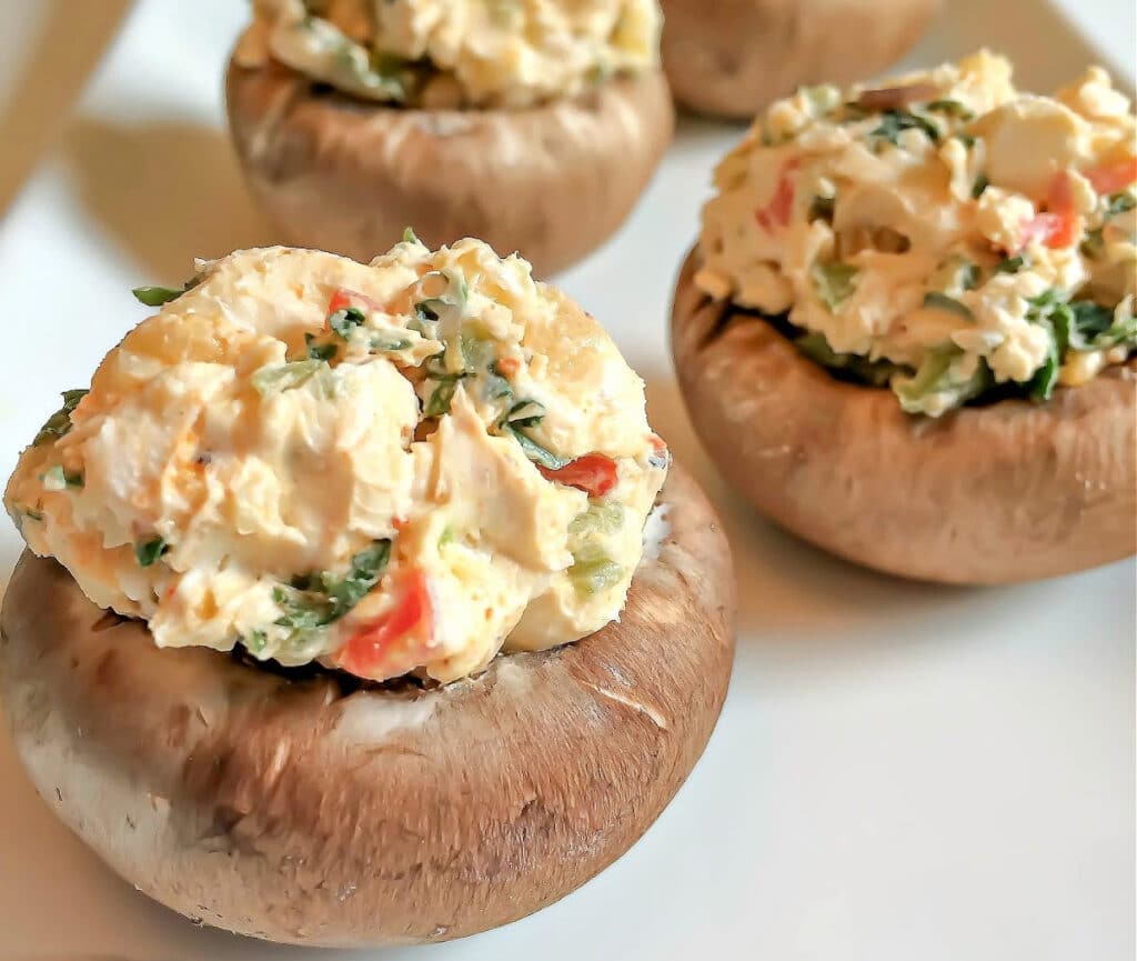 Close-up of four Cajun Spiced Stuffed Mushrooms on a white plate, filled with a creamy mixture of chopped vegetables and herbs. The colorful filling has visible green and red bits, adding a bold kick to each bite.