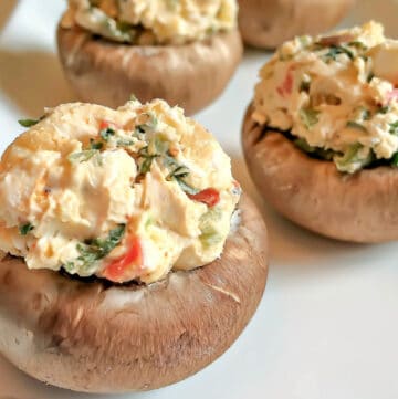 Close-up of four Cajun Spiced Stuffed Mushrooms on a white plate, filled with a creamy mixture of chopped vegetables and herbs. The colorful filling has visible green and red bits, adding a bold kick to each bite.