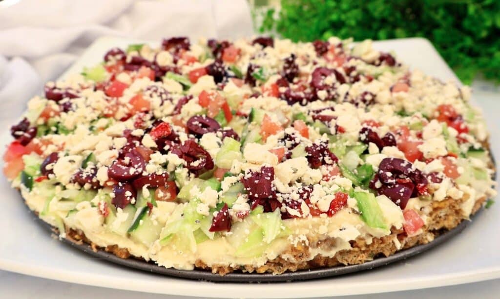 A close-up of a baked falafel tart layered with chopped lettuce, tomatoes, kalamata olives, and crumbled feta cheese, displayed on a round platter with greenery in the background.