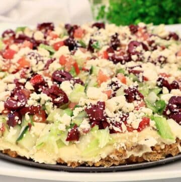 A close-up of a baked falafel tart layered with chopped lettuce, tomatoes, kalamata olives, and crumbled feta cheese, displayed on a round platter with greenery in the background.
