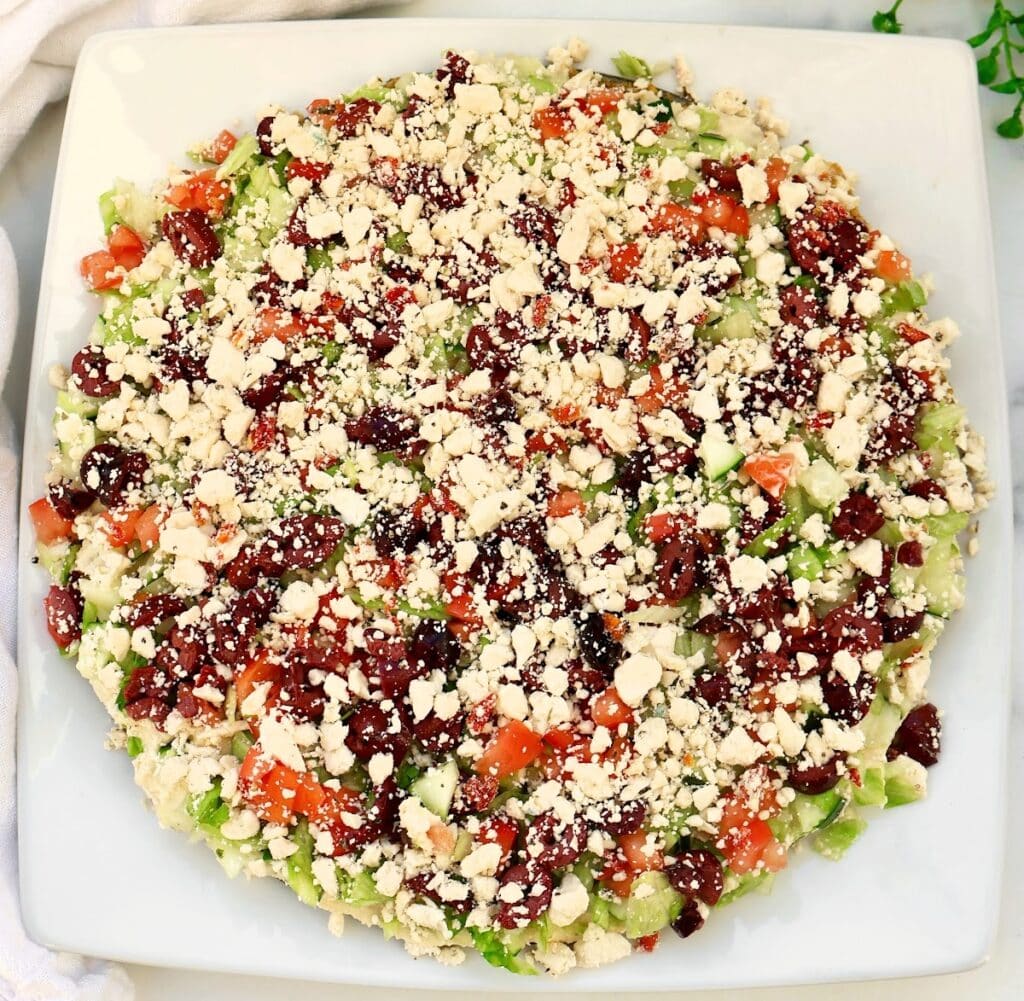 A baked falafel pie on a square white plate, topped with crumbled cheese, diced tomatoes, cucumbers, green peppers, onions, and olives, arranged in a colorful pattern.