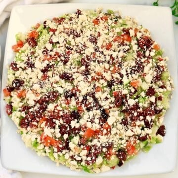 A baked falafel pie on a square white plate, topped with crumbled cheese, diced tomatoes, cucumbers, green peppers, onions, and olives, arranged in a colorful pattern.