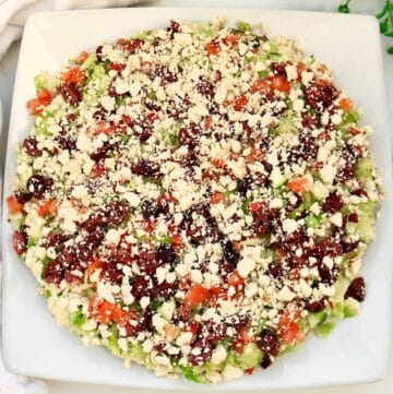 A baked falafel pie on a square white plate, topped with crumbled cheese, diced tomatoes, cucumbers, green peppers, onions, and olives, arranged in a colorful pattern.