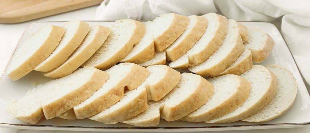 A rectangular white plate with several slices of French bread baguette arranged in neat rows. A wooden cutting board and a white cloth are visible in the background, showcasing this easy, quick serving idea.