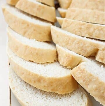 Slices of fresh white bread, similar to a classic French bread baguette, are arranged in overlapping rows on a white surface. The bread has a soft crumb and a light golden crust, making it an easy and quick choice for any meal.