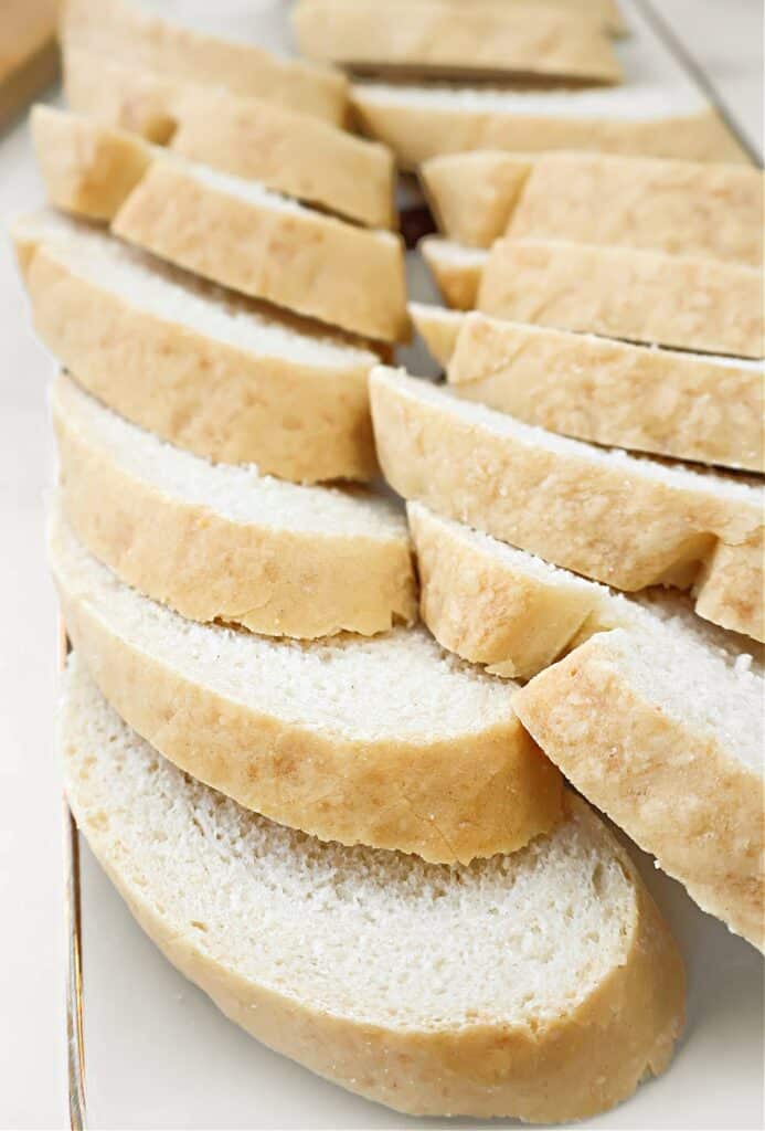 Slices of fresh white bread, similar to a classic French bread baguette, are arranged in overlapping rows on a white surface. The bread has a soft crumb and a light golden crust, making it an easy and quick choice for any meal.
