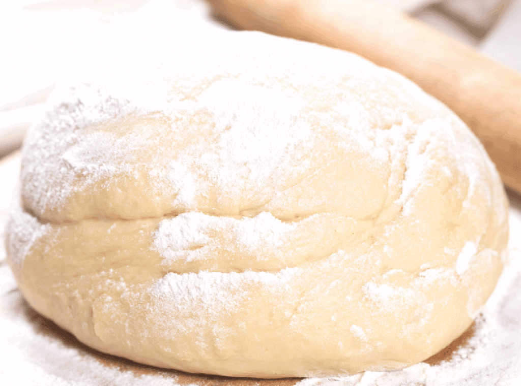 A ball of homemade pizza dough dusted with flour sits on a lightly floured surface, with a wooden rolling pin blurred in the background.