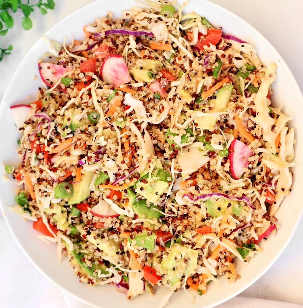 A white bowl filled with a healthy quinoa salad featuring shredded cabbage, carrots, radishes, avocado, green onions, and red bell peppers, all garnished with fresh herbs.