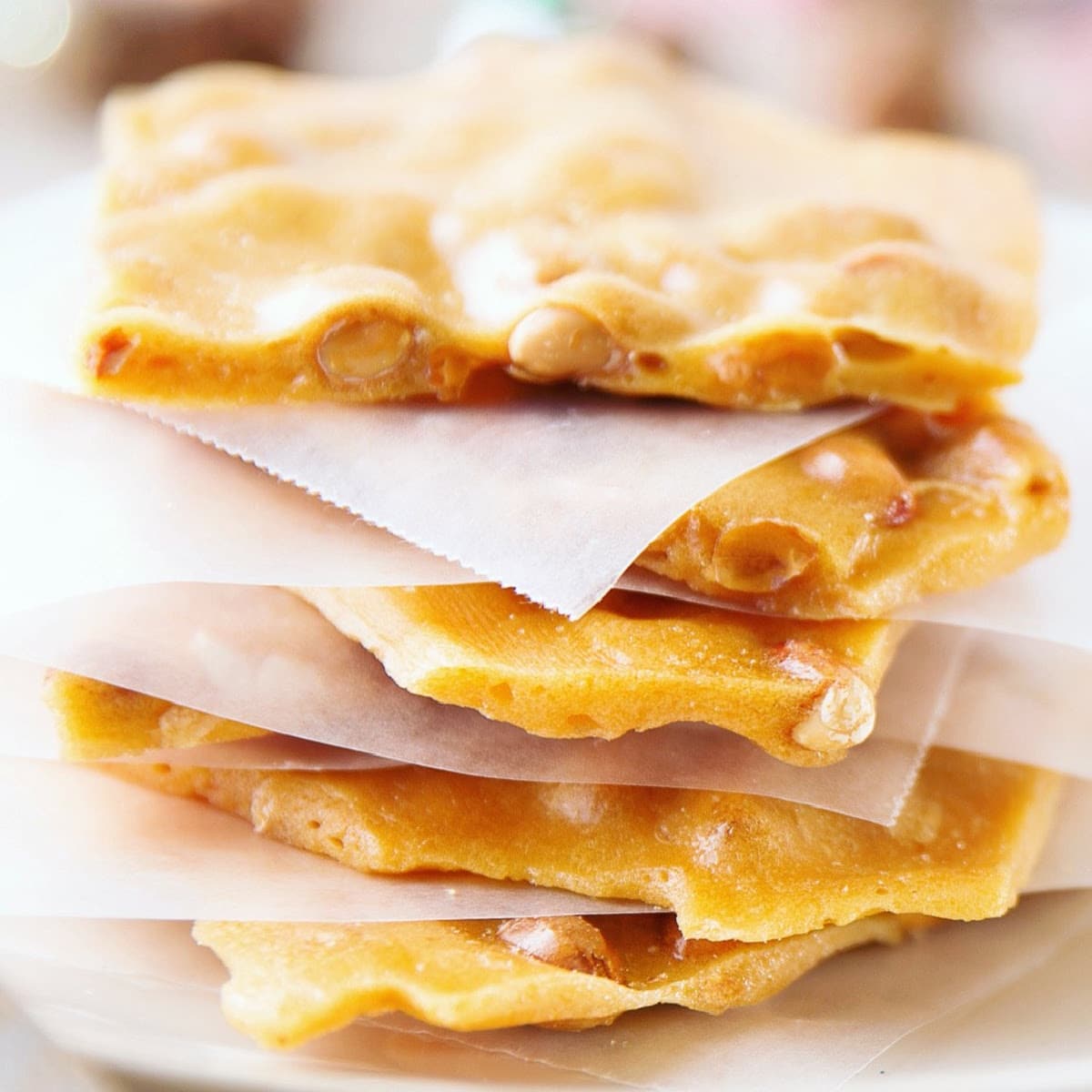 A close-up of several stacked pieces of traditional vegan peanut brittle separated by sheets of wax paper, showing the golden, glossy candy embedded with peanuts.