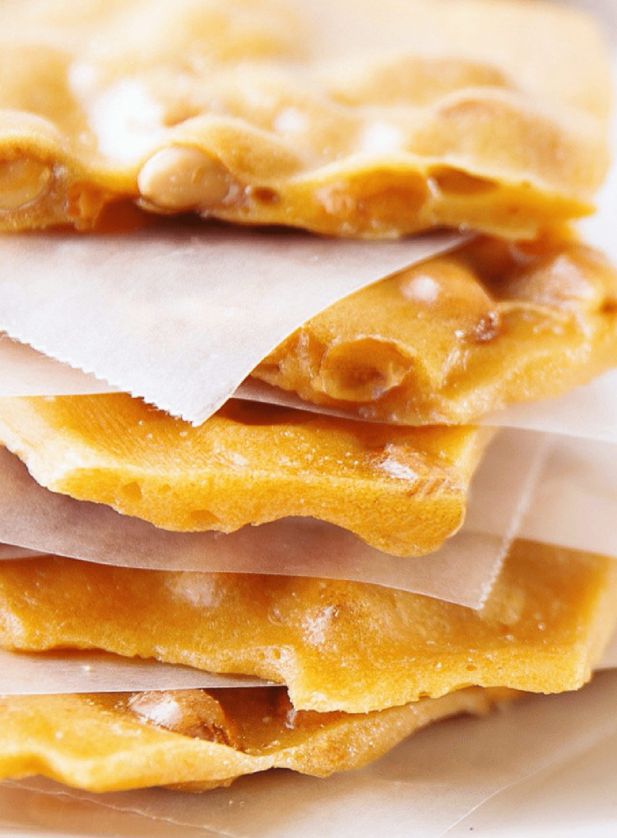 Close-up of stacked pieces of traditional vegan peanut brittle separated by sheets of wax paper, highlighting the glossy, crunchy texture and visible peanuts embedded in the candy.