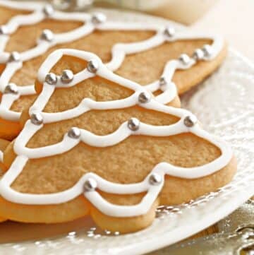 Christmas tree-shaped sugar cookies with white icing and silver ball decorations are arranged on a white plate, featuring vegan royal icing for a festive and plant-based treat.