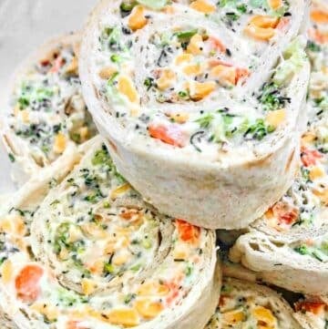 Vegan veggie pinwheels made from tortillas rolled with a creamy filling of chopped broccoli, carrots, cheese, and herbs, sliced into spiral rounds and neatly stacked on a plate.