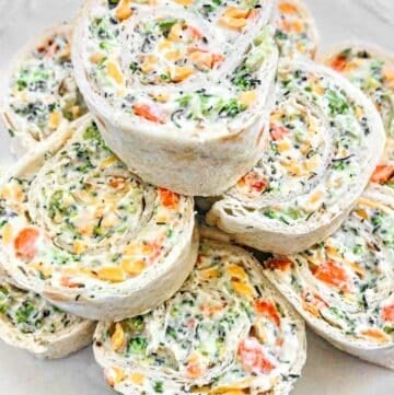 A plate of vegan veggie pinwheels made with tortillas filled with cream cheese, shredded carrots, broccoli, and other vegetables. The pinwheels are neatly stacked and display vibrant orange and green hues.