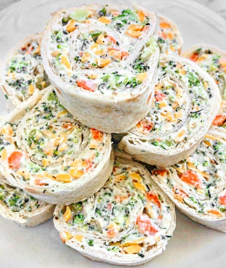 A plate of vegan veggie pinwheels made with tortillas filled with cream cheese, shredded carrots, broccoli, and other vegetables. The pinwheels are neatly stacked and display vibrant orange and green hues.
