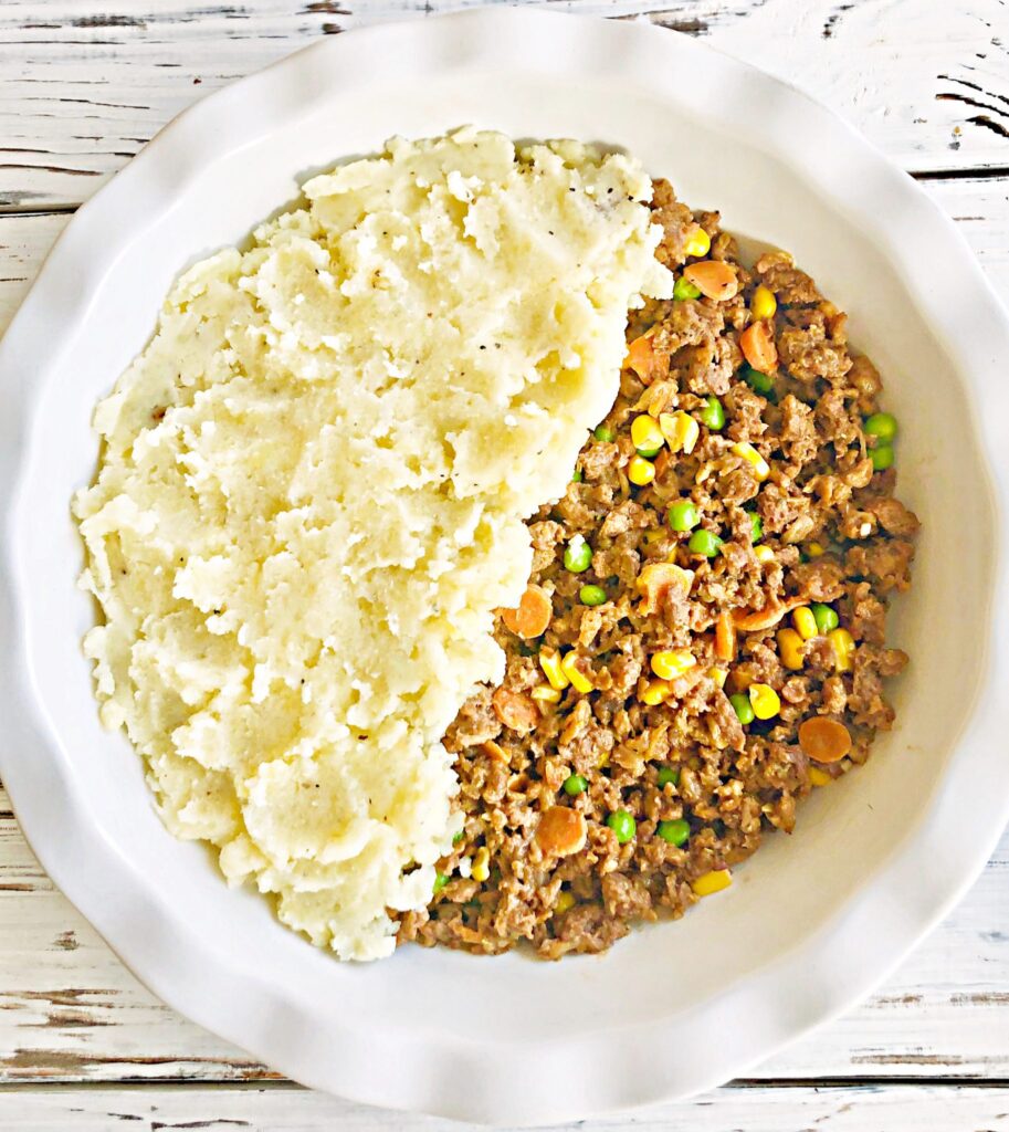 A round white dish containing mashed potatoes on one half and a vegan cottage pie mixture with peas, corn, and carrots on the other half, placed on a white wooden surface.