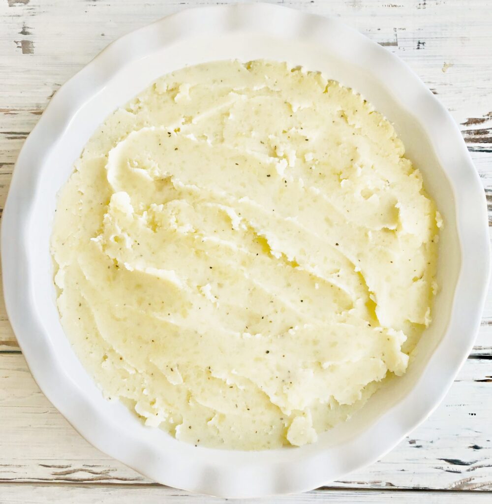 A white ceramic dish filled with smooth, creamy mashed potatoes—perfect for topping a vegan cottage pie—sits on a rustic white wooden surface, its surface lightly flecked with black pepper.