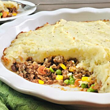 A close-up of a vegan cottage pie in a white baking dish, with a portion removed to reveal hearty vegetables under mashed potatoes; a serving is on a plate in the background.