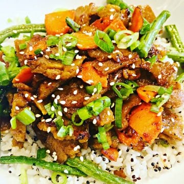 A close-up of a seitan rice bowl filled with white rice, topped with stir-fried vegetables, sliced carrots, green beans, and plant-based meat, garnished with sliced green onions and sesame seeds.