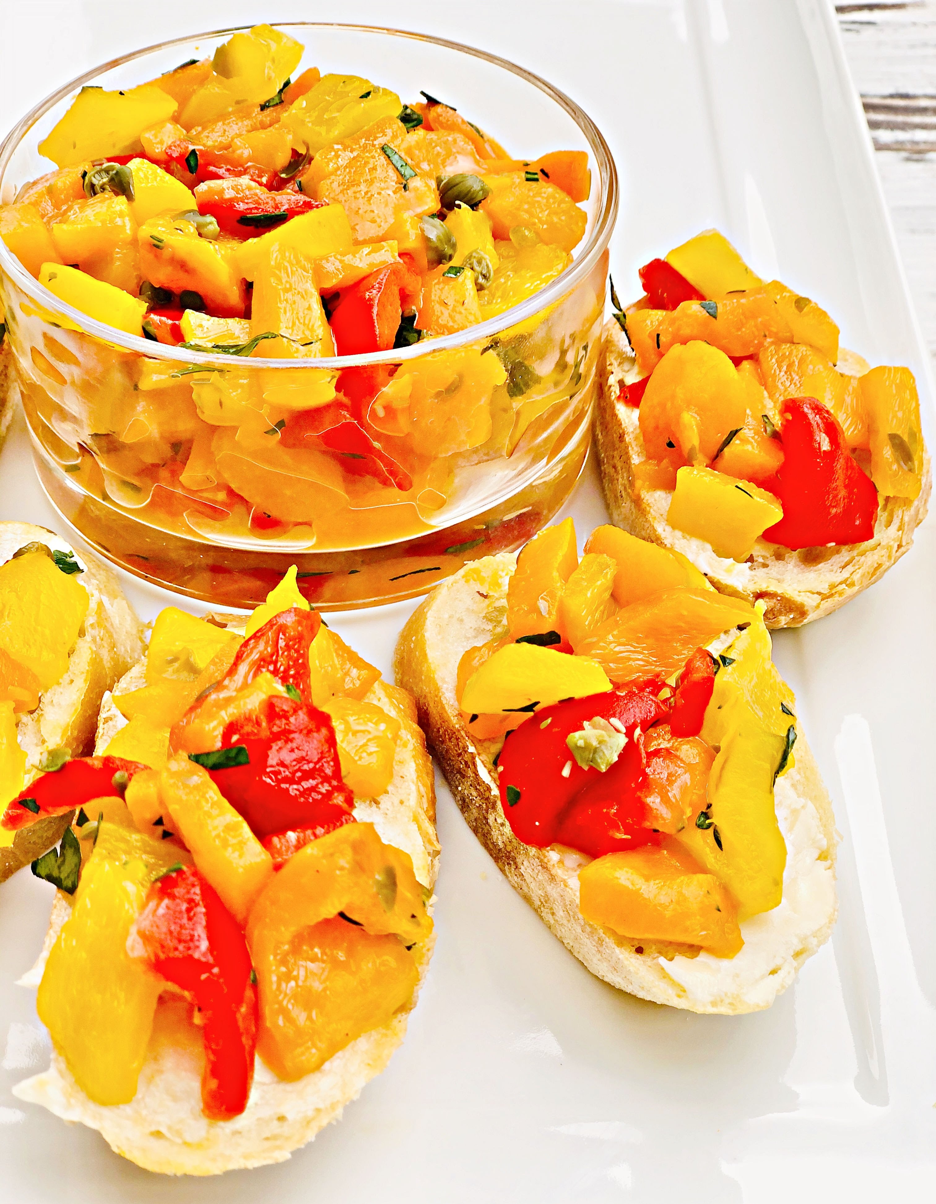 Slices of bread topped with vibrant bell pepper bruschetta and fresh herbs, served alongside a glass bowl of the same roasted pepper mixture on a white plate.