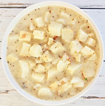 A white bowl filled with creamy baked potato soup, featuring chunks of potatoes and specks of seasoning, placed on a white wooden surface.