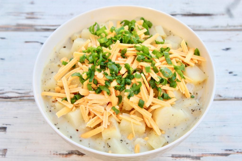 A white bowl filled with creamy baked potato soup, topped with shredded cheddar cheese and chopped green onions, set on a rustic white wooden surface.