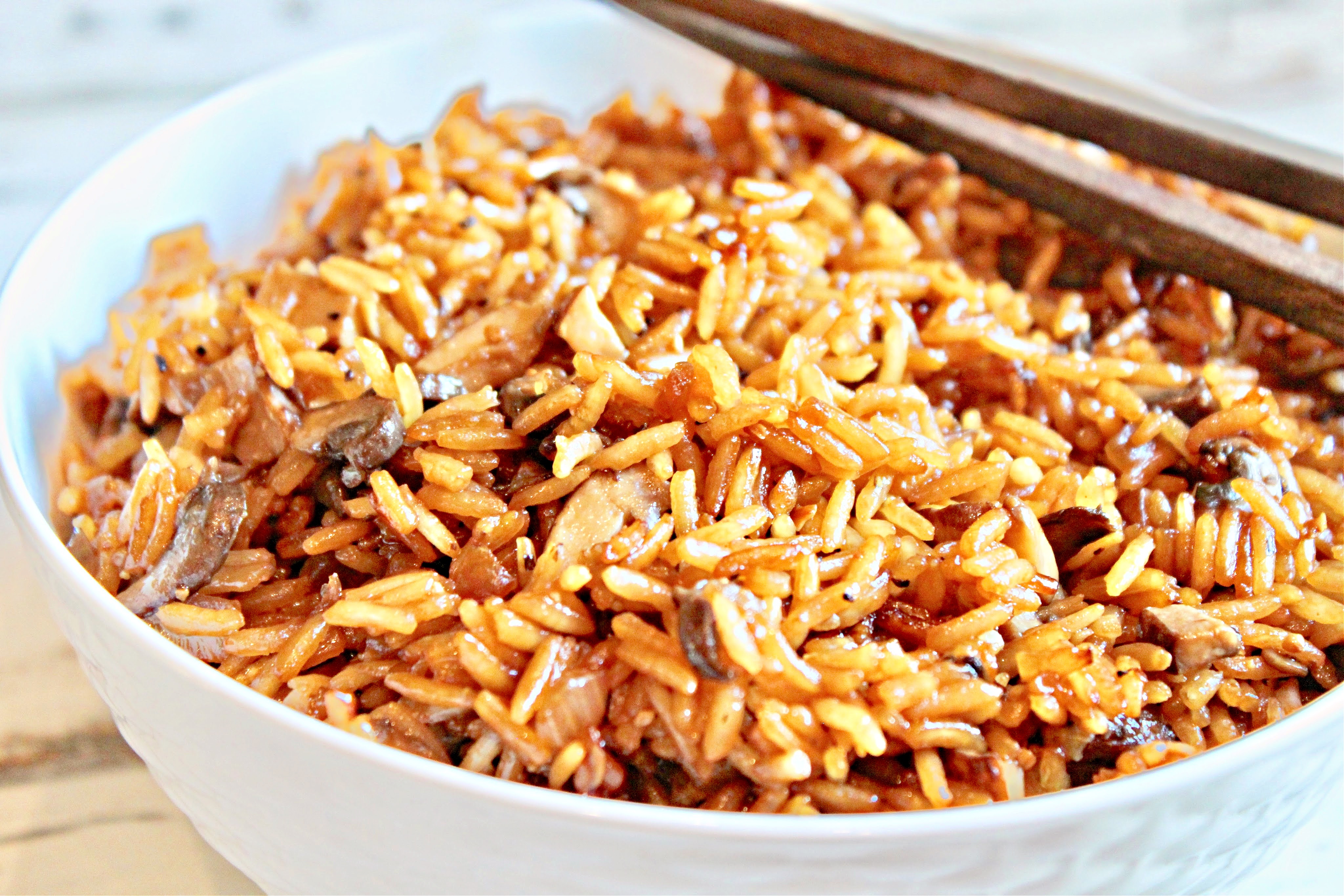 A close-up of a white bowl filled with asian mushroom rice mixed with soy sauce, with chopsticks resting on the rim of the bowl.