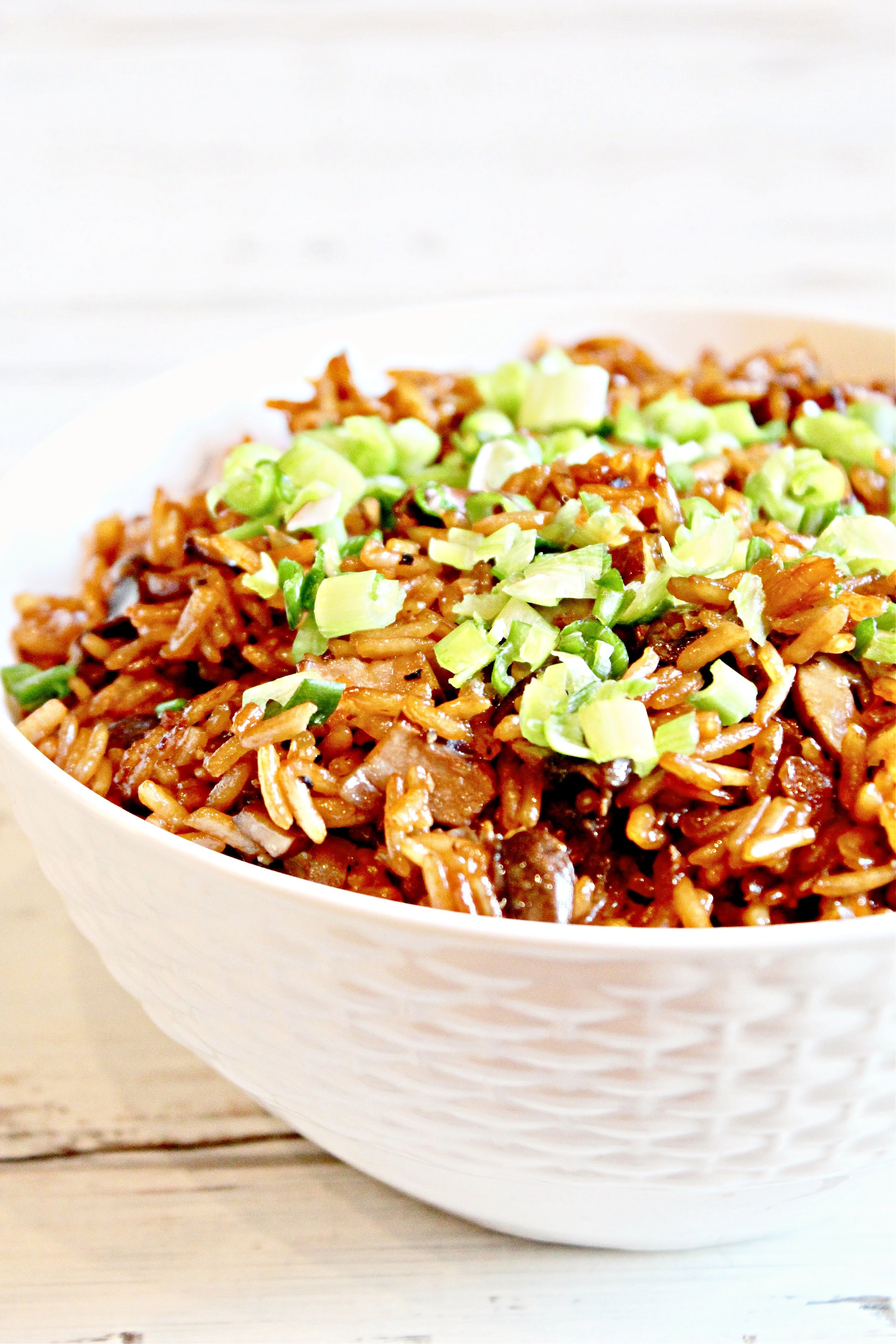 A white bowl filled with asian mushroom rice mixed with vegetables and topped with chopped green onions, set on a light wooden surface.