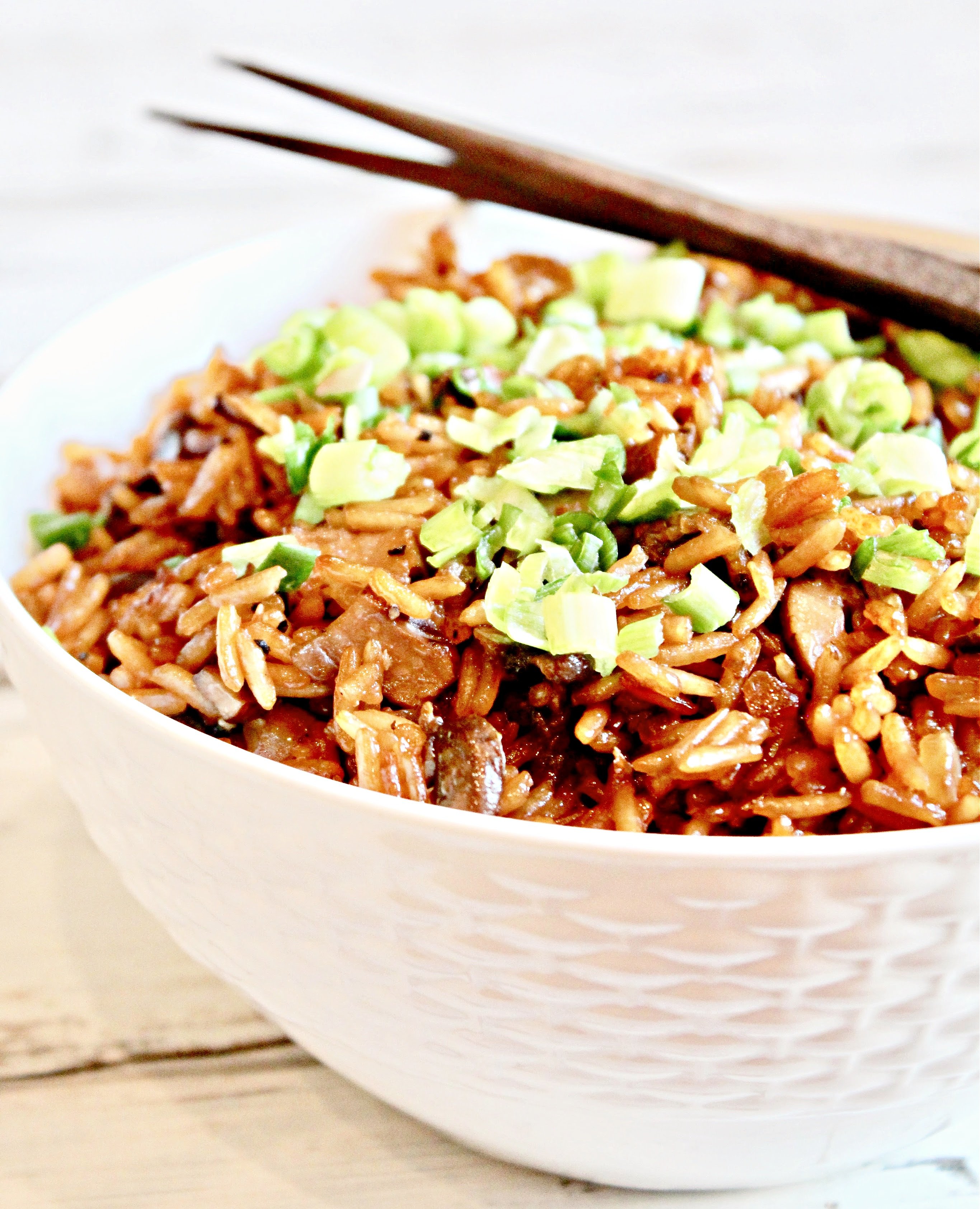 A white bowl filled with asian mushroom rice topped with chopped green onions, with a pair of wooden chopsticks resting on the edge of the bowl.