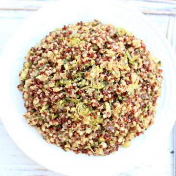 A white plate filled with a colorful quinoa salad, featuring red and white quinoa mixed with cheesy broccoli quinoa and finely shredded green vegetables, set on a light wooden surface.