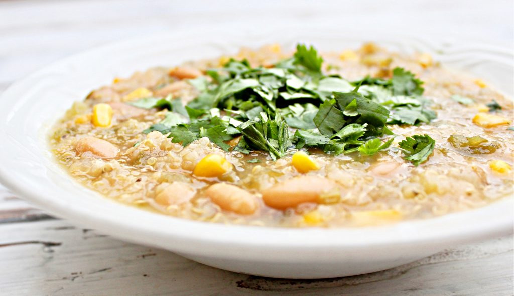 A white bowl filled with a hearty white bean quinoa chili, featuring beans, corn, and quinoa, topped with a generous amount of fresh chopped cilantro, on a light wooden surface.