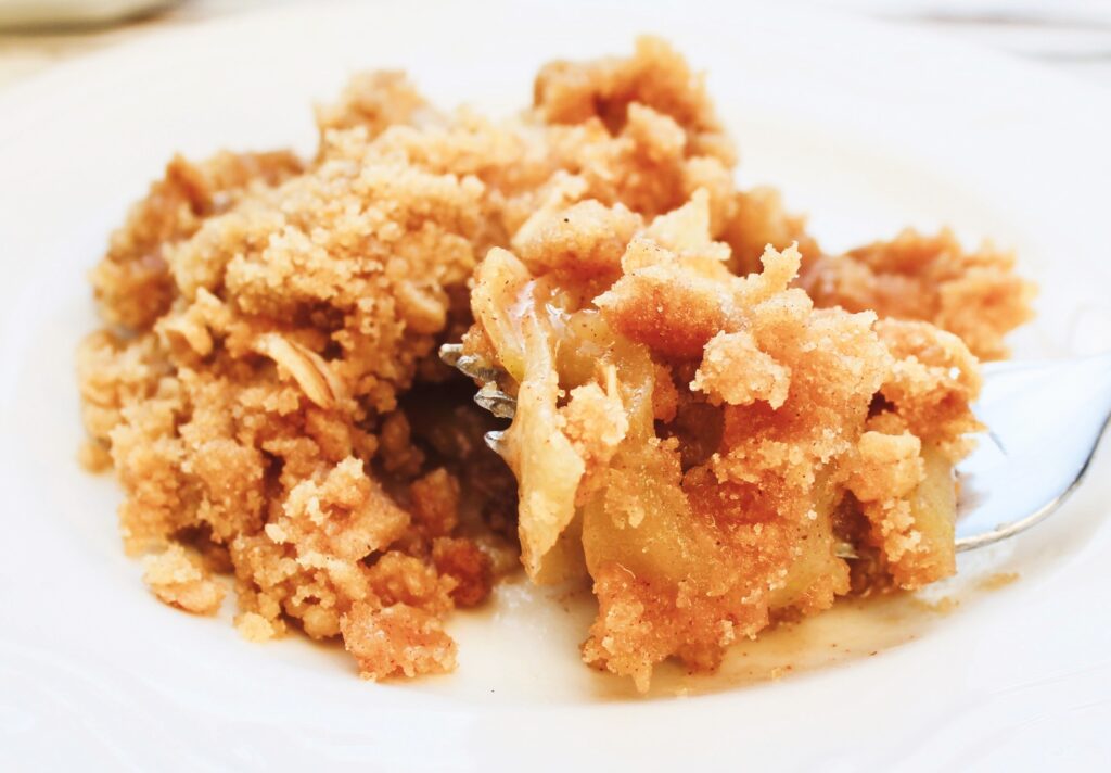 A close-up of a serving of apple crisp, featuring a golden, crumbly topping and pieces of baked apple, on a white plate with a fork.