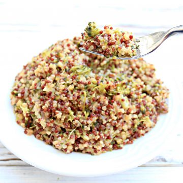 A close-up of a plate filled with cheesy broccoli quinoa mixed with other vegetables. A fork lifts a portion above the plate, highlighting the colorful, cheesy quinoa grains and vibrant vegetable pieces.