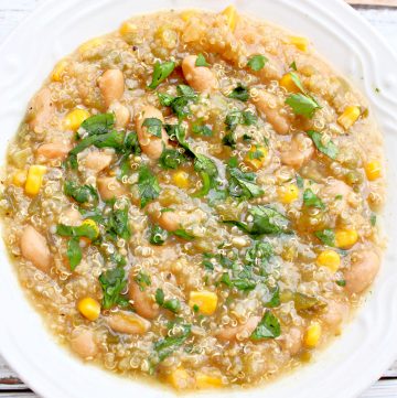 A white bowl filled with creamy white bean quinoa chili, featuring corn, quinoa, and chopped fresh cilantro on top. The dish has a hearty, textured appearance with a light, greenish broth.