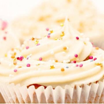 Close-up of a cupcake with swirled vanilla buttercream frosting, decorated with colorful sprinkles and small stars, in a white paper liner. Other cupcakes are softly blurred in the background.