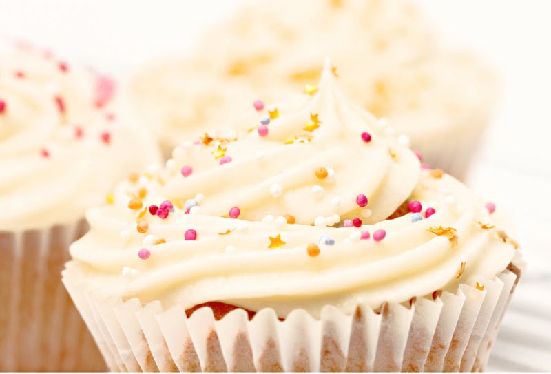 Close-up of a cupcake with creamy white vanilla buttercream frosting, topped with colorful round sprinkles and small gold star sprinkles, in a white paper wrapper. Other similarly decorated cupcakes are blurred in the background.