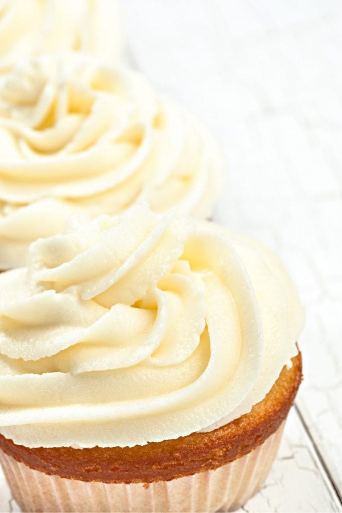 Close-up of vanilla cupcakes topped with swirls of creamy vanilla buttercream frosting, set on a light textured surface.