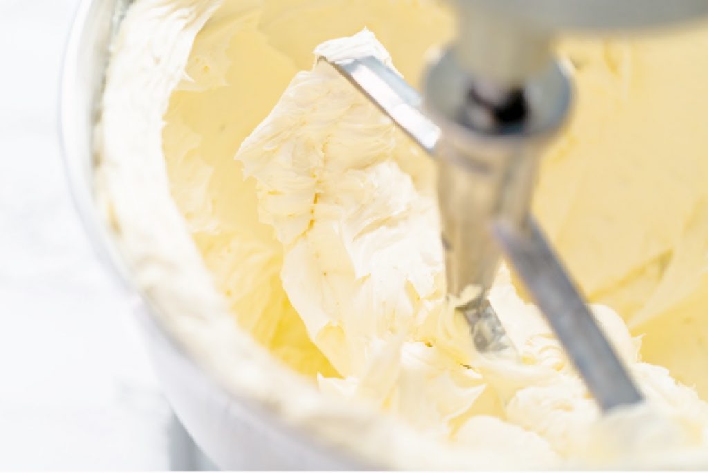 Close-up of creamy, whipped vanilla buttercream in a mixing bowl with a metal paddle attachment from a stand mixer. The texture appears smooth and fluffy, with soft peaks forming on the surface.